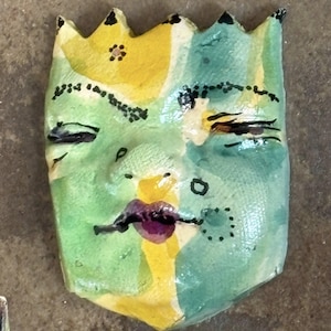May include: Handmade ceramic mask with a crown-like top. The face is painted with green, yellow, and blue hues, featuring closed eyes, a purple mouth, and black details. The mask has a textured surface and a unique, artistic design.