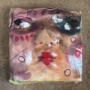 May include: A square, handmade ceramic tile featuring a painted face. The face has black eyes, red lips, and various colorful accents, including purple, red, and green. The tile has a rough, textured surface and a unique, artistic style.