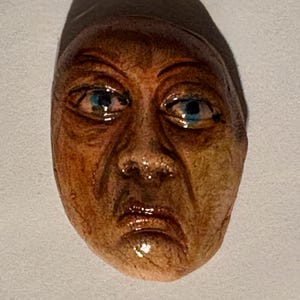 May include: A sculpted face ornament featuring a textured, weathered skin tone with deep brown shading, prominent facial lines, and bright blue eyes. The figure has a downward-turned mouth and a dark, smooth, rounded cap covering the top of the head.
