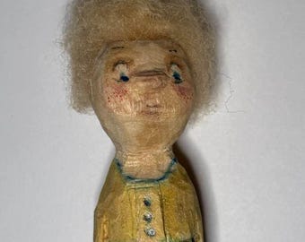 Könnte beinhalten: Handgeschnitzte Holzpuppe mit hellem Teint, blonden Haaren und blauen Augen. Die Puppe trägt ein gelbes Oberteil mit drei kleinen Knöpfen und einen grün-gelb gestreiften Rock. Die Puppe hat einen leicht traurigen Ausdruck.