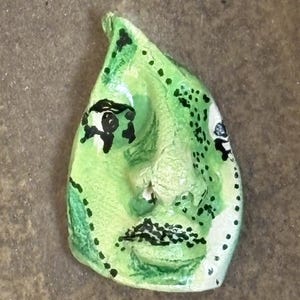 May include: A handmade, abstract face sculpture with a light green base color. The face features black details for the eyes, eyebrows, and mouth. The sculpture has a textured surface with black dotted accents and white highlights.