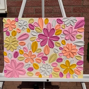 May include: A rectangular canvas with raised floral designs in pink, yellow, orange, and white. The flowers and petals are arranged on a gradient background, creating a textured, three-dimensional effect. The artwork is displayed on a white easel.