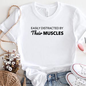 May include: White t-shirt with the text "Easily Distracted By Their Muscles" printed in black.