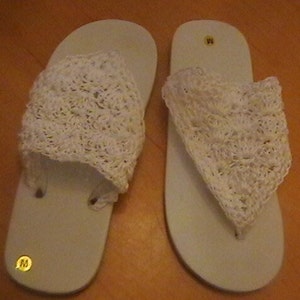 Könnte beinhalten: Ein Paar weiße Flip-Flops mit gehäkelten Riemen. Die Riemen sind weiß und haben ein zartes, spitzenartiges Muster. Die Flip-Flops haben ein weißes 'W'-Etikett am Riemen.