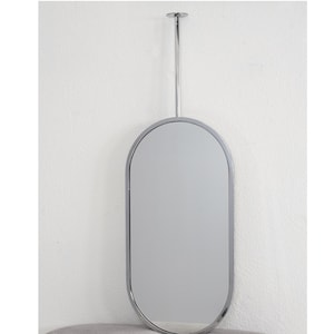Puede incluir: Un espejo ovalado con un marco de metal pulido y una varilla metálica larga y delgada para colgarlo. El espejo está montado en una pared blanca, reflejando el entorno. El diseño es minimalista y moderno, ideal para cualquier espacio.