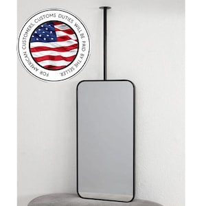 Pode incluir: Um espelho retangular com cantos arredondados, emoldurado em preto, suspenso por uma haste preta fixada ao teto. O espelho é exibido contra uma parede branca. O texto "FOR AMERICAN CUSTOMERS, CUSTOMS DUTIES WILL BE PAID BY THE SELLER" está em um círculo.