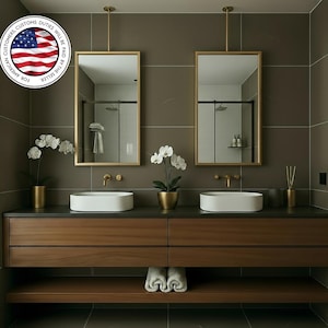 Pode incluir: Um design de casa de banho moderno com dois espelhos retangulares com moldura dourada suspensos sobre uma bancada de madeira escura com duas pias brancas. O espaço inclui torneiras douradas, orquídeas e uma prateleira com toalhas dobradas. As paredes são cinza neutro.