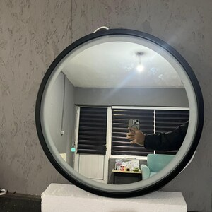 Puede incluir: Un espejo de pared circular con un marco negro elegante y una tira de luz LED integrada a lo largo del borde interior. El espejo est&aacute; colocado sobre un bloque de espuma blanco rectangular, reflejando el interior de una habitaci&oacute;n.