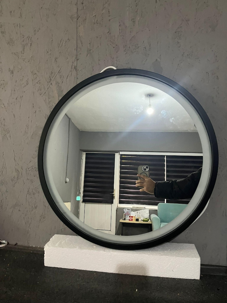 Puede incluir: Un espejo de pared circular con un marco negro elegante y una tira de luz LED integrada a lo largo del borde interior. El espejo est&aacute; colocado sobre un bloque de espuma blanco rectangular, reflejando el interior de una habitaci&oacute;n.