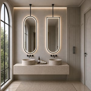 May include: Two wall-mounted mirrors with black frames and integrated LED lighting. One mirror is oval, the other has a flat top with rounded bottom corners. Both hang from black ceiling rods above two beige stone vessel sinks on a floating vanity.