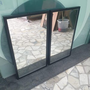 May include: Two rectangular mirrors with black frames are placed next to each other. The mirrors reflect the surroundings, including a light-coloured floor and various objects. The frames have a simple, modern design.