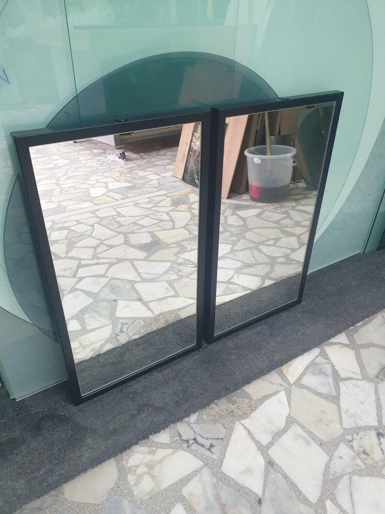 May include: Two rectangular mirrors with black frames are placed next to each other. The mirrors reflect the surroundings, including a light-coloured floor and various objects. The frames have a simple, modern design.