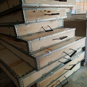 May include: A pile of rectangular wooden crates with metal reinforced edges and black handles. The crates are made of light-coloured wood and are stacked. The metal edges are silver.