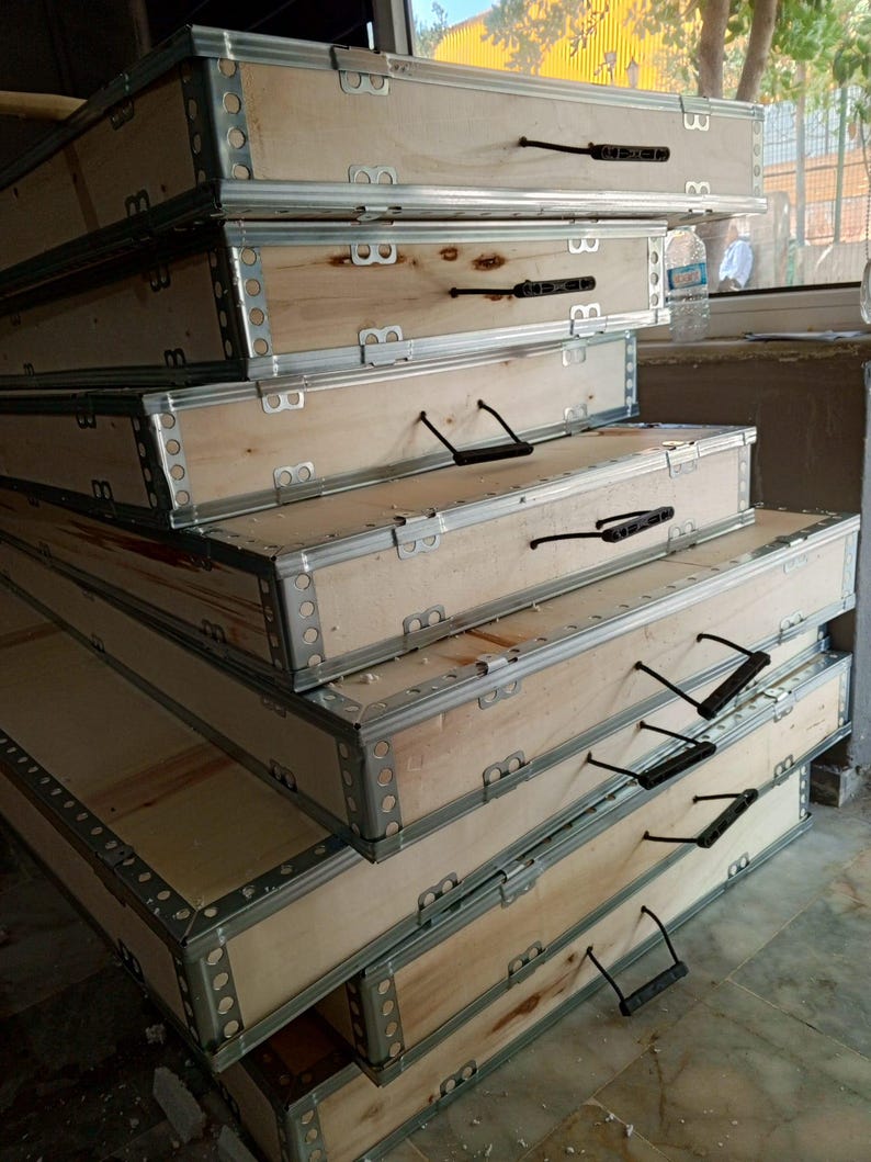 May include: A pile of rectangular wooden crates with metal reinforced edges and black handles. The crates are made of light-coloured wood and are stacked. The metal edges are silver.
