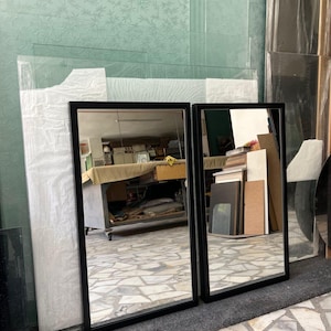 May include: Two rectangular mirrors with black frames. The mirrors reflect a room with various objects. The mirrors are leaning against a wall with a light green floral pattern. The floor is covered with a gray rug and a patterned tile.