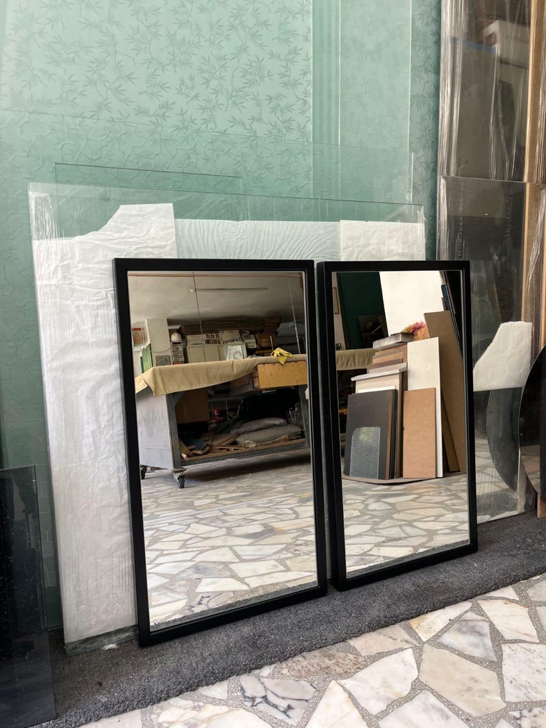 May include: Two rectangular mirrors with black frames. The mirrors reflect a room with various objects. The mirrors are leaning against a wall with a light green floral pattern. The floor is covered with a gray rug and a patterned tile.