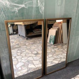 May include: Two rectangular mirrors with gold-coloured frames. The mirrors are leaning against a wall, reflecting the surroundings. The frames have a simple, modern design.