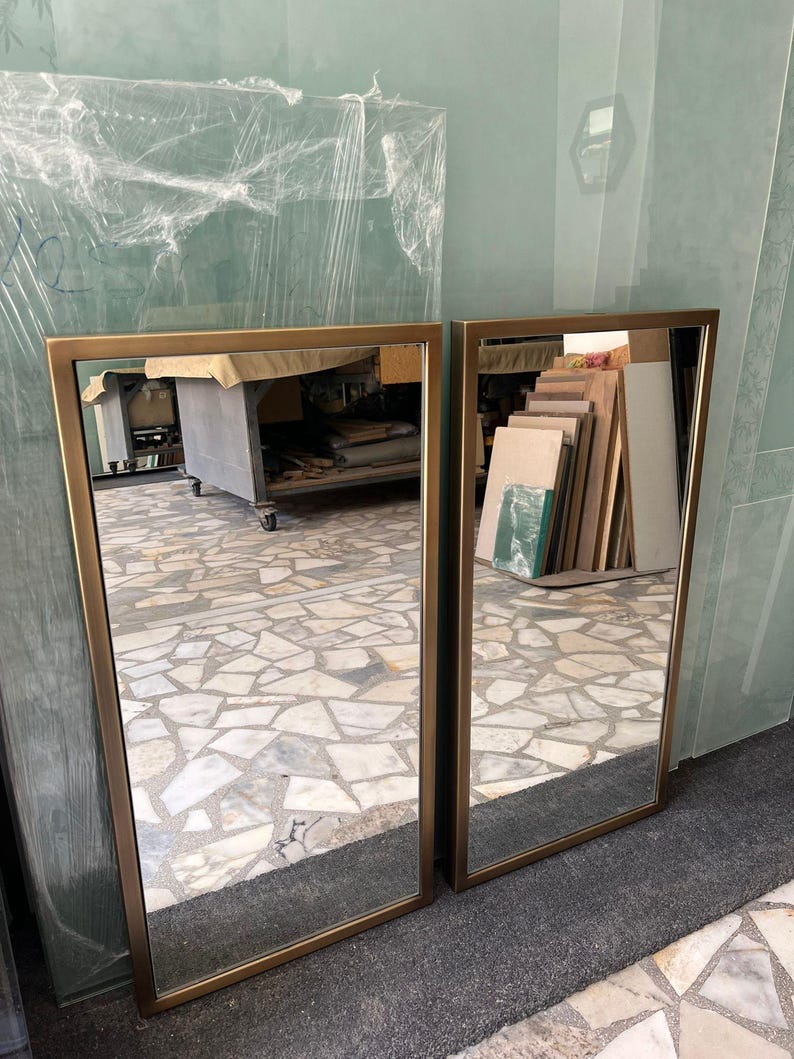 May include: Two rectangular mirrors with gold-coloured frames. The mirrors are leaning against a wall, reflecting the surroundings. The frames have a simple, modern design.