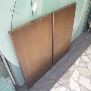 May include: Two bronze-colored, rectangular panels with a trapezoidal shape. Each panel has a hole near the top and is secured with small rivets. The panels are leaning against a glass surface, suggesting a possible architectural or design element.