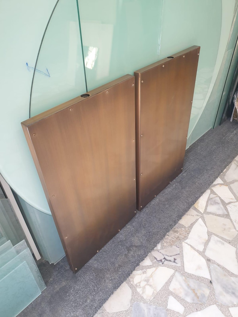 May include: Two bronze-colored, rectangular panels with a trapezoidal shape. Each panel has a hole near the top and is secured with small rivets. The panels are leaning against a glass surface, suggesting a possible architectural or design element.