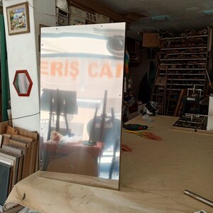 May include: A large, rectangular mirror with a reflective surface stands on a table covered with a tan cloth. The mirror reflects the interior of a workshop, including tables, tools, and the text "ERIS CAT".