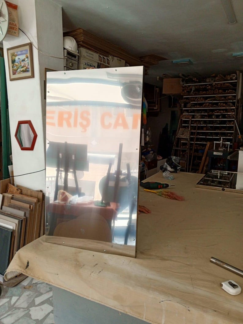 May include: A large, rectangular mirror with a reflective surface stands on a table covered with a tan cloth. The mirror reflects the interior of a workshop, including tables, tools, and the text "ERIS CAT".