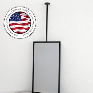 May include: A rectangular black framed mirror suspended from a black rod attached to the ceiling. The mirror is positioned against a white wall. A circular graphic with the American flag and text is in the upper left corner.
