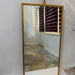 May include: A rectangular floor mirror featuring a metallic gold frame. The mirror is suspended from a matching gold-colored cylindrical ceiling mount that extends from the top center of the frame, creating a sleek and vertical display arrangement.