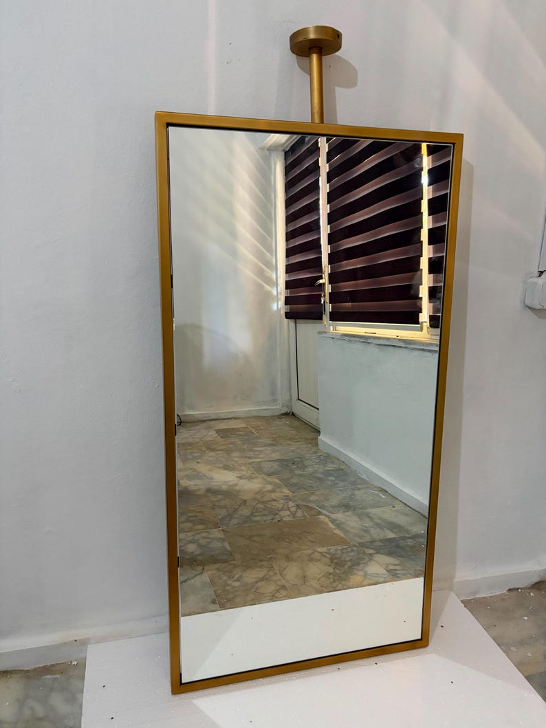 May include: A rectangular floor mirror featuring a metallic gold frame. The mirror is suspended from a matching gold-colored cylindrical ceiling mount that extends from the top center of the frame, creating a sleek and vertical display arrangement.