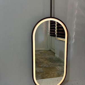 Puede incluir: Un espejo de pared de forma ovalada con un marco de color marr&oacute;n oscuro y una tira de luz LED integrada a lo largo del borde interior. El espejo est&aacute; montado sobre una varilla vertical que se extiende hacia arriba, ofreciendo un dise&ntilde;o elegante.