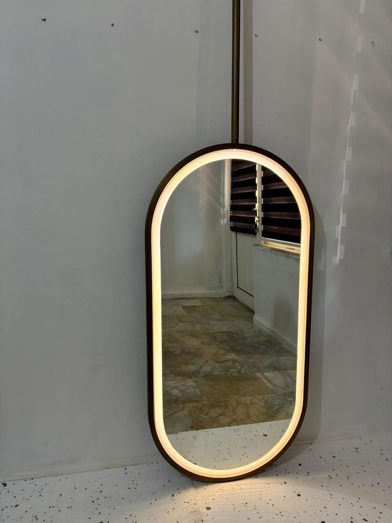 Puede incluir: Un espejo de pared de forma ovalada con un marco de color marr&oacute;n oscuro y una tira de luz LED integrada a lo largo del borde interior. El espejo est&aacute; montado sobre una varilla vertical que se extiende hacia arriba, ofreciendo un dise&ntilde;o elegante.