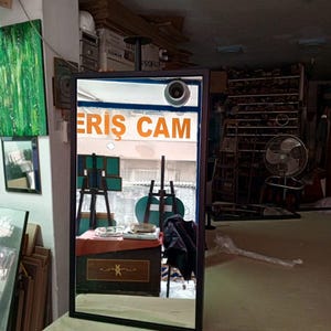 May include: A black-framed rectangular mirror reflecting a shop interior. The mirror shows a table, easels, and the orange text "ERIS CAM". The mirror is placed on a light-coloured surface.