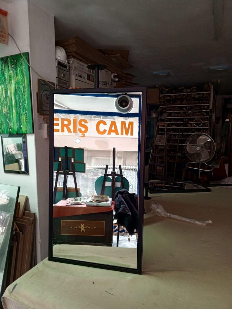 May include: A black-framed rectangular mirror reflecting a shop interior. The mirror shows a table, easels, and the orange text "ERIS CAM". The mirror is placed on a light-coloured surface.