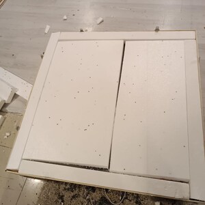 May include: A partially assembled white cabinet door with a rectangular frame. The door is made of white foam and has a visible seam down the middle. The frame is made of wood. The cabinet is on a light-coloured floor.