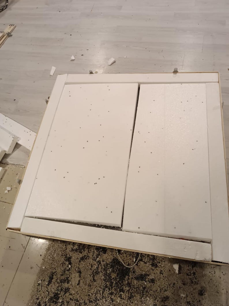 May include: A partially assembled white cabinet door with a rectangular frame. The door is made of white foam and has a visible seam down the middle. The frame is made of wood. The cabinet is on a light-coloured floor.