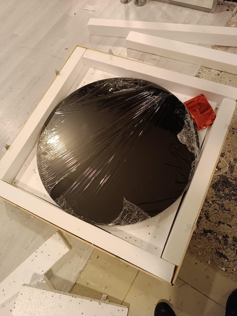 Puede incluir: Un objeto circular negro con acabado brillante, parcialmente envuelto en film pl&aacute;stico transparente, dentro de una caja de env&iacute;o cuadrada de cart&oacute;n blanco con inserciones de espuma protectora y un peque&ntilde;o paquete rojo rectangular.