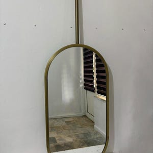 Puede incluir: Un espejo de forma ovalada con un marco met&aacute;lico en tono lat&oacute;n. El espejo est&aacute; suspendido de una varilla vertical que se fija a una placa de montaje circular en la parte superior, permitiendo que cuelgue del techo o de una superficie.