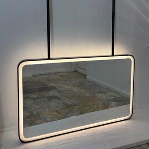 Puede incluir: Un espejo de pared rectangular con marco negro e iluminaci&oacute;n LED integrada a lo largo del borde interior. El espejo est&aacute; suspendido por dos varillas verticales negras fijadas a la parte superior del marco, emitiendo un suave brillo blanco.