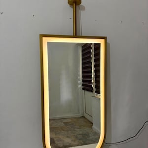 Puede incluir: Un espejo de pared rectangular con el borde inferior redondeado y un marco de metal dorado. El espejo cuenta con una tira de luz LED integrada a lo largo del per&iacute;metro interior del marco. Una varilla vertical dorada conecta la parte superior con un soporte circular.