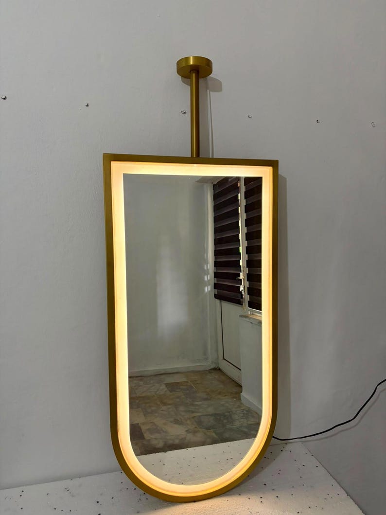 Puede incluir: Un espejo de pared rectangular con el borde inferior redondeado y un marco de metal dorado. El espejo cuenta con una tira de luz LED integrada a lo largo del per&iacute;metro interior del marco. Una varilla vertical dorada conecta la parte superior con un soporte circular.