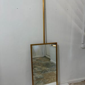 May include: A rectangular mirror with a metallic gold frame, suspended from a vertical gold rod that attaches to a circular ceiling mount. The mirror features a clean, straight-edged design with a reflective glass surface and a matching gold finish.