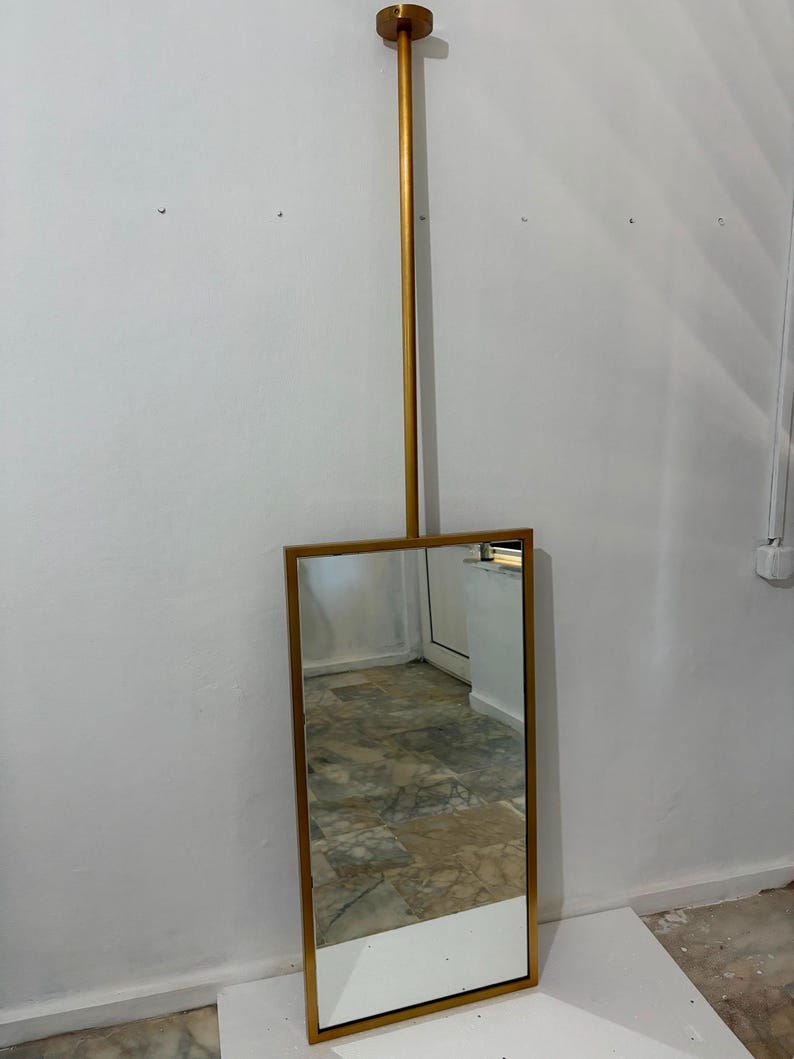 May include: A rectangular mirror with a metallic gold frame, suspended from a vertical gold rod that attaches to a circular ceiling mount. The mirror features a clean, straight-edged design with a reflective glass surface and a matching gold finish.