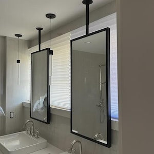 May include: Rectangular bathroom mirrors with black frames hanging from the ceiling. The mirrors reflect the bathroom's interior, including a shower and sinks. The design is modern and minimalist, with clean lines and a focus on functionality.