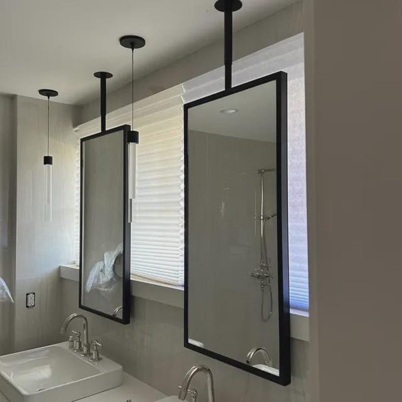 May include: Rectangular bathroom mirrors with black frames hanging from the ceiling. The mirrors reflect the bathroom's interior, including a shower and sinks. The design is modern and minimalist, with clean lines and a focus on functionality.