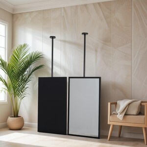 May include: Two rectangular mirrors, one black and one reflective, suspended from the ceiling by black rods. The mirrors are in a room with a neutral colour palette, a palm tree, and a wooden bench.