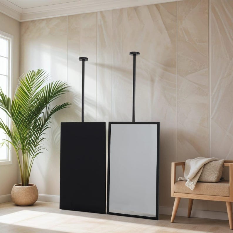 May include: Two rectangular mirrors, one black and one reflective, suspended from the ceiling by black rods. The mirrors are in a room with a neutral colour palette, a palm tree, and a wooden bench.