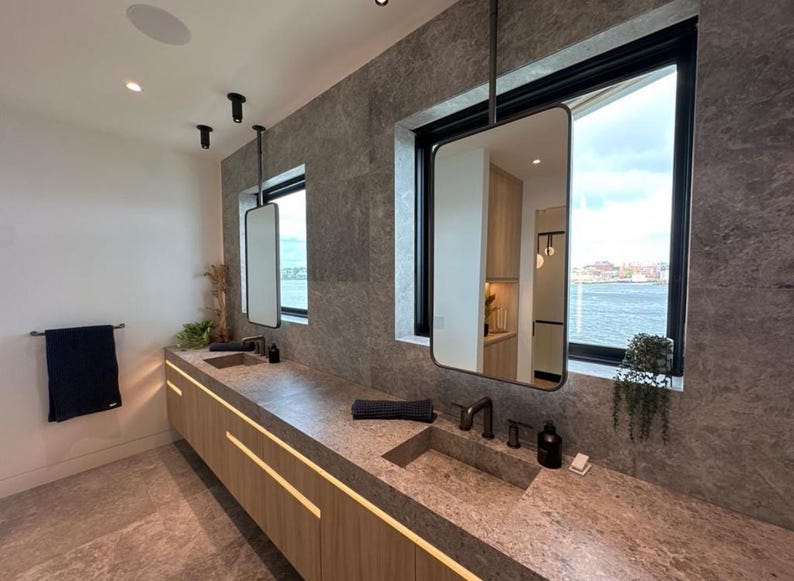 K&ouml;nnte beinhalten: Ein modernes Badezimmer mit zwei Waschbecken, einem gro&szlig;en Fenster mit Blick auf das Wasser und einem schwebenden Waschtisch mit Holzschr&auml;nken.