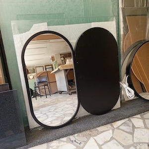 Puede incluir: Un espejo de doble panel con una secci&oacute;n de cristal ovalada reflectante con marco negro y un panel ovalado negro s&oacute;lido. El lado reflectante muestra el interior de un taller, mientras que un cable blanco sale del panel negro.