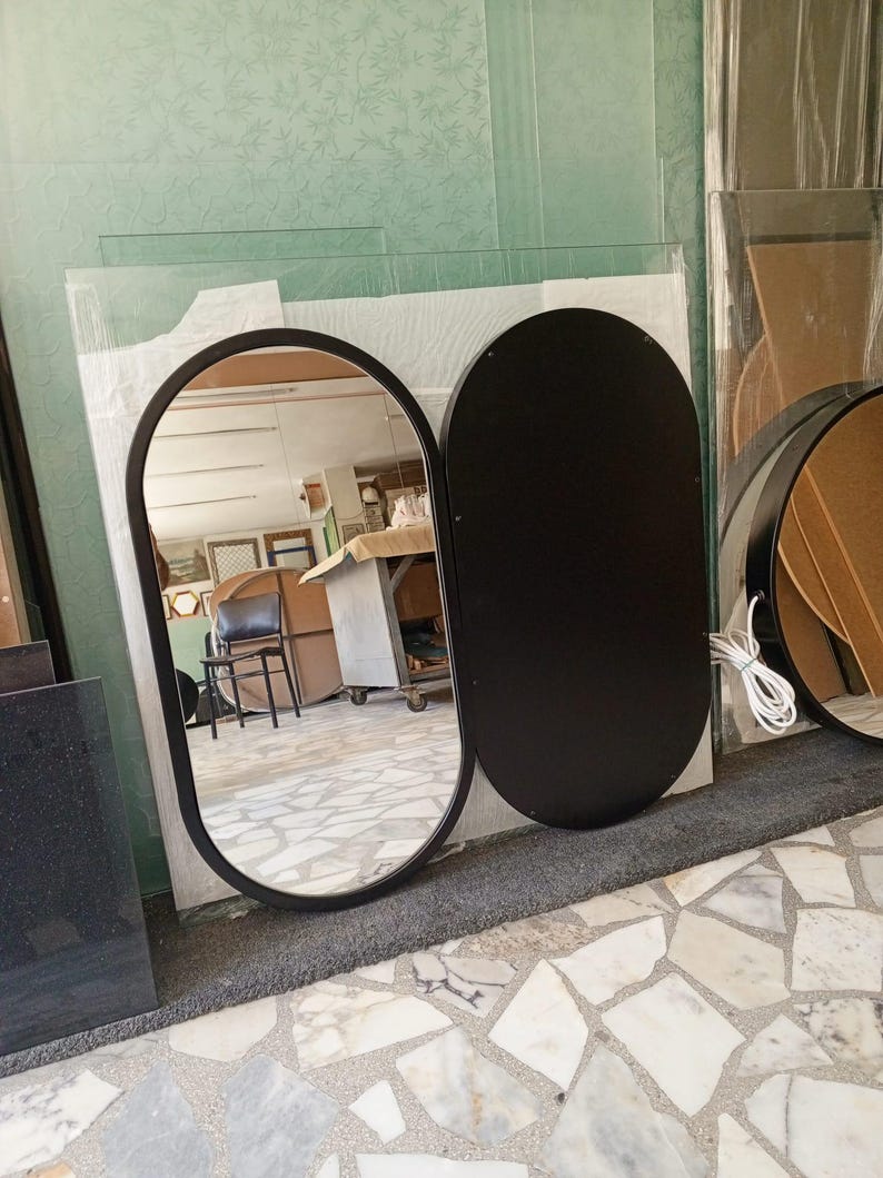 Puede incluir: Un espejo de doble panel con una secci&oacute;n de cristal ovalada reflectante con marco negro y un panel ovalado negro s&oacute;lido. El lado reflectante muestra el interior de un taller, mientras que un cable blanco sale del panel negro.