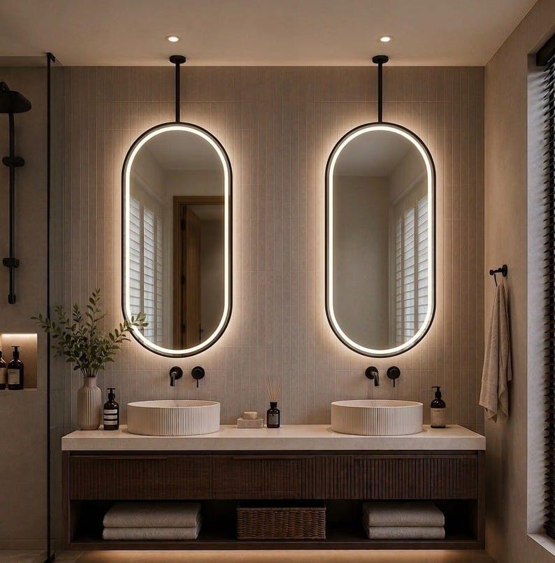 Puede incluir: Dos espejos de pared ovalados con retroiluminaci&oacute;n LED integrada, suspendidos del techo mediante varillas de metal negro. Los espejos se sit&uacute;an sobre un mueble de ba&ntilde;o doble con dos lavabos redondos de cer&aacute;mica estriada.